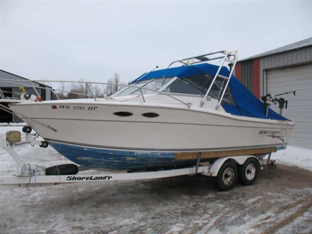 White boat with blue cover mounted on a ShoreLand'r trailer equipped with an Atlantic Towers Arch beside a building.