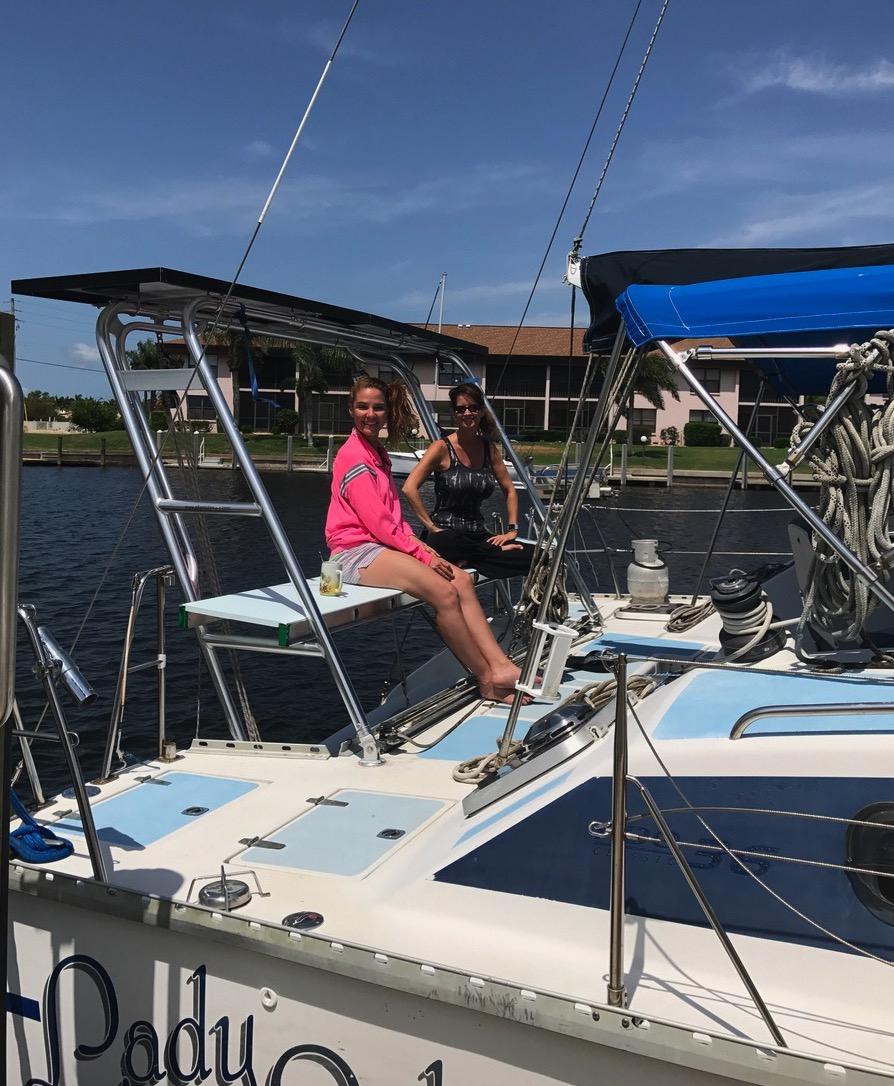 Two women sitting on a sailboat equipped with Atlantic Towers Arch, docked near waterfront buildings on a clear day.
