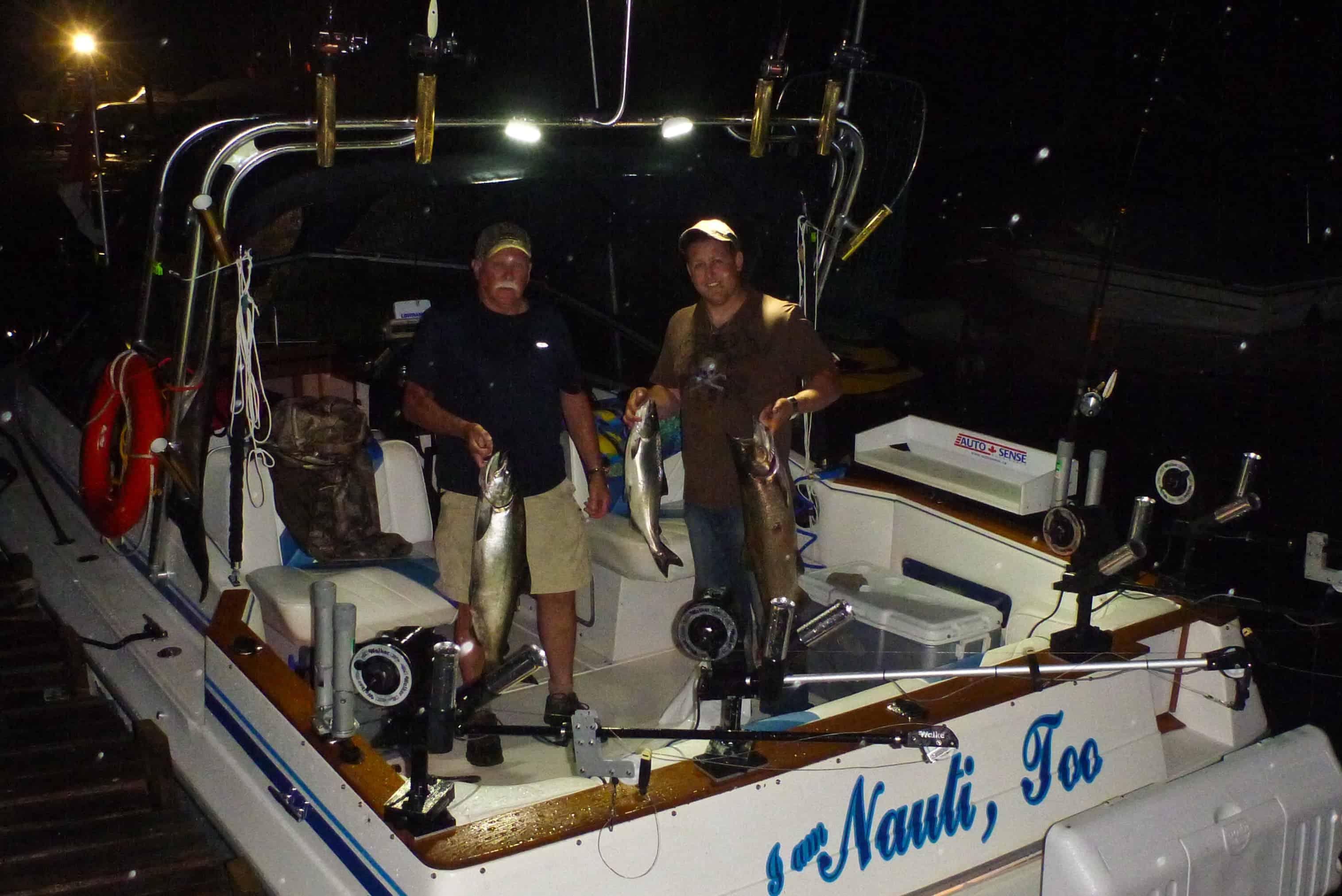 Two men standing on a fishing boat at night holding large fish they caught, with fishing gear visible on the boat.