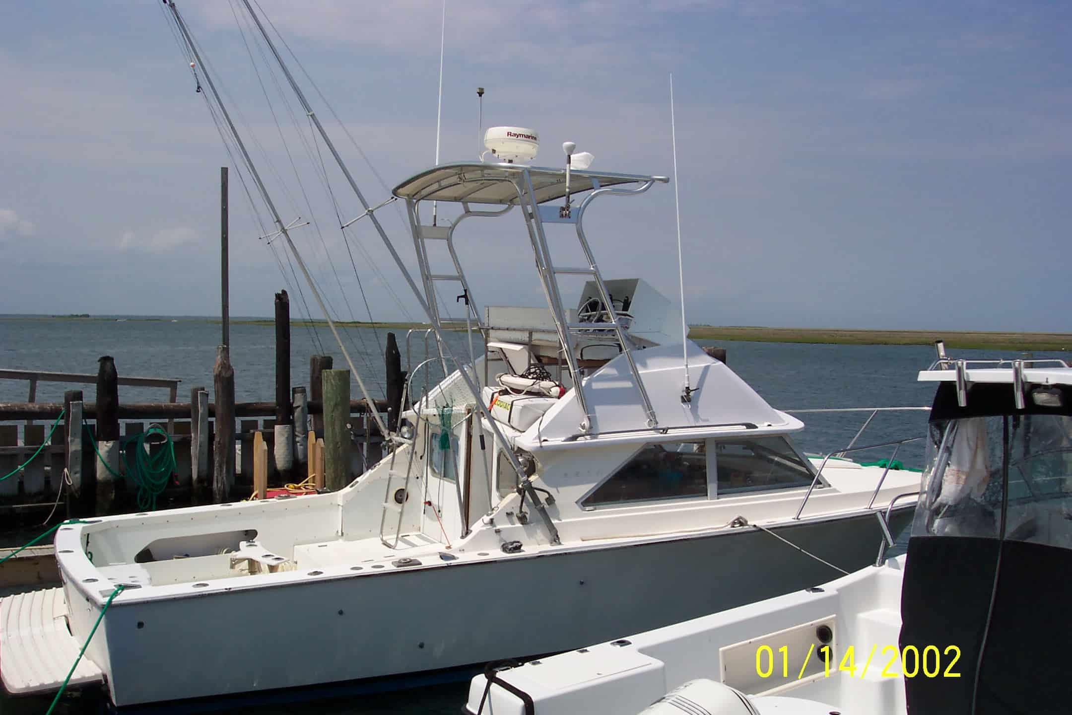 White fishing boat docked by a wooden pier with an Atlantic Towers arch and radar dome under a partly cloudy sky.