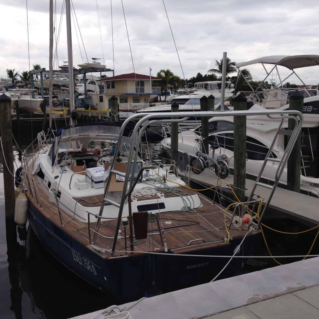 Sailboat with wooden deck docked at marina pier under overcast sky, surrounded by other moored boats and palm trees.