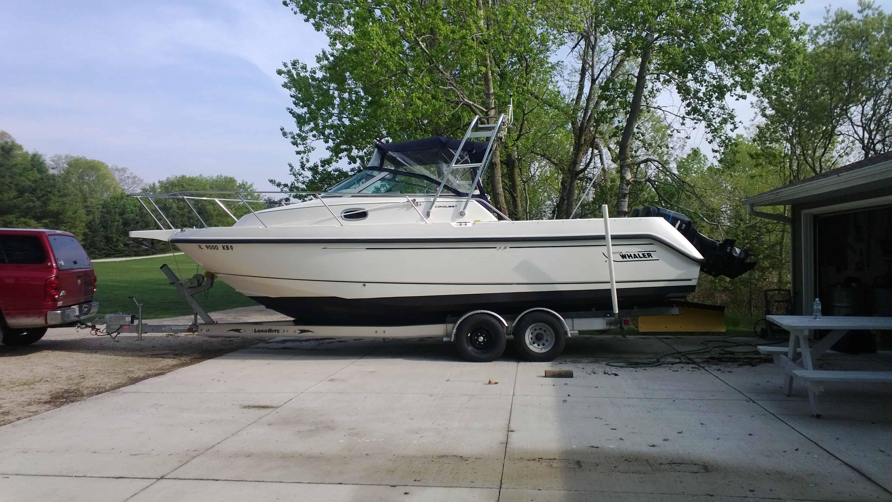 Side view of a white boat on a trailer hitched to a red truck, parked on a driveway near a garage and trees in the background.