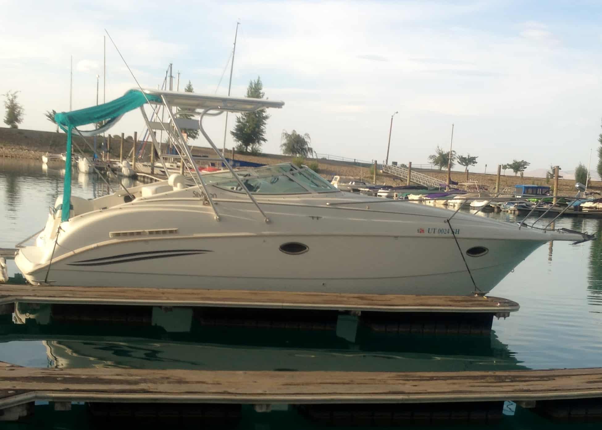 White motorboat docked at a marina with a green fabric canopy and fishing rods on a calm water background.