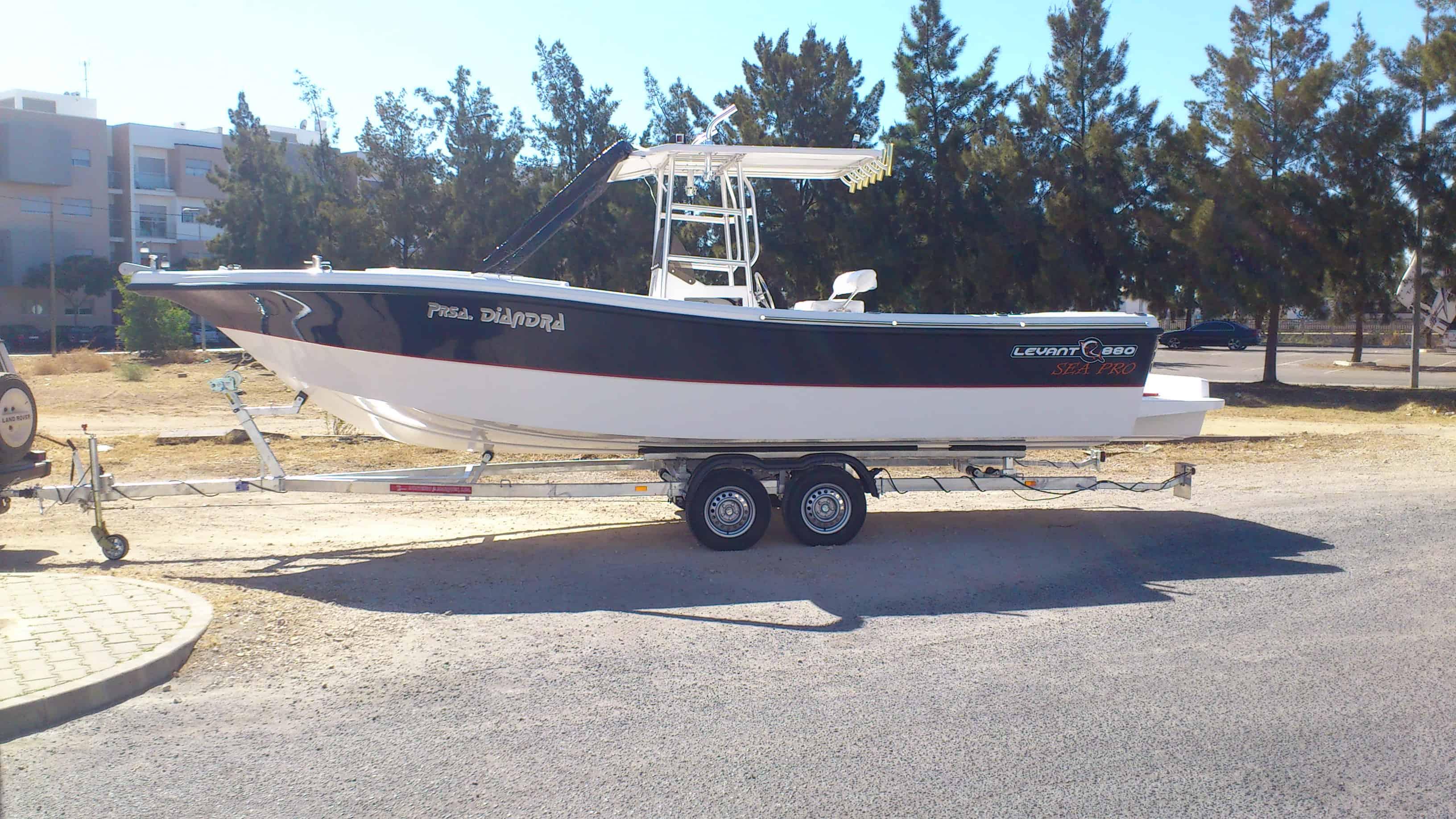 Side view of a black and white boat with center console on a dual-axle trailer parked on a paved road with trees and buildings in the background.