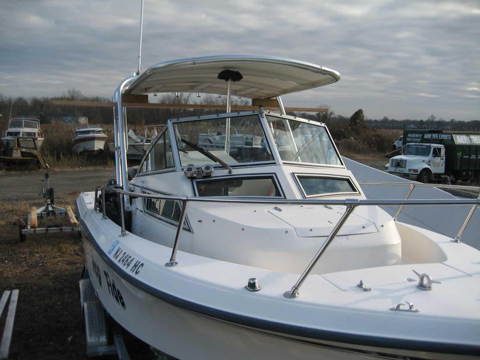 White boat with windshield and covered cabin area, parked on trailer in a lot with other boats and vehicles in background under cloudy sky