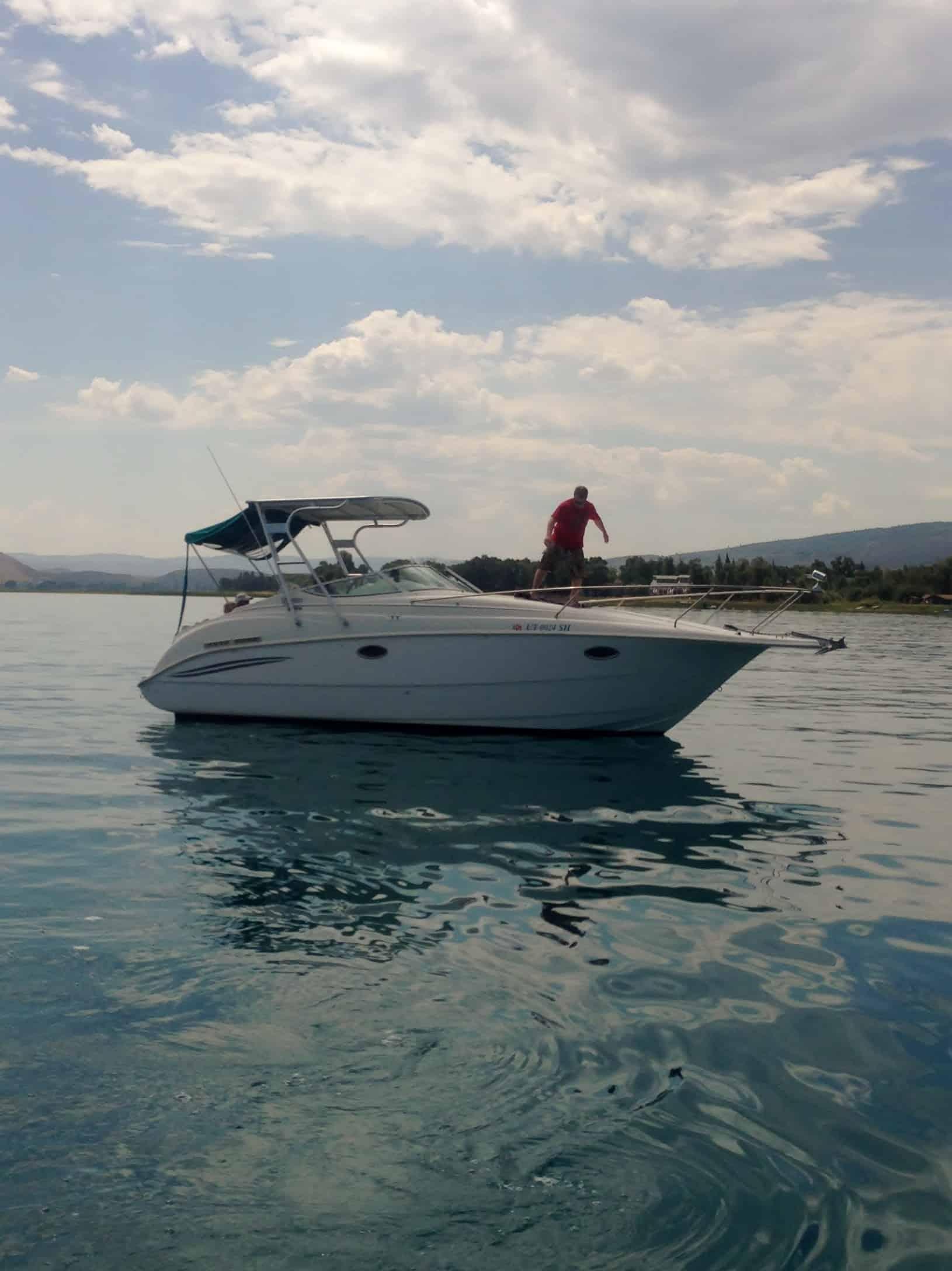 A person standing on the bow of a small white motorboat floating on calm water with hills and cloudy sky in the background.