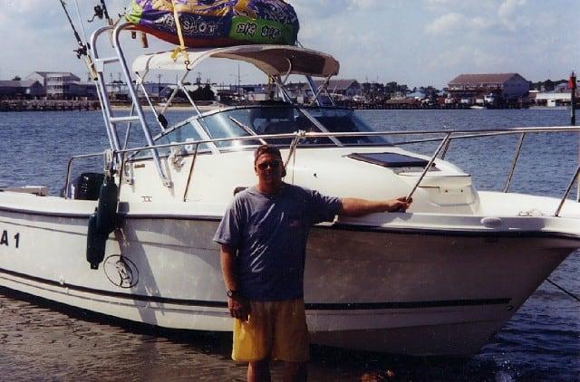 Man standing beside a white 2302 Trophy boat equipped with an Atlantic Towers Arch on calm water near a marina.