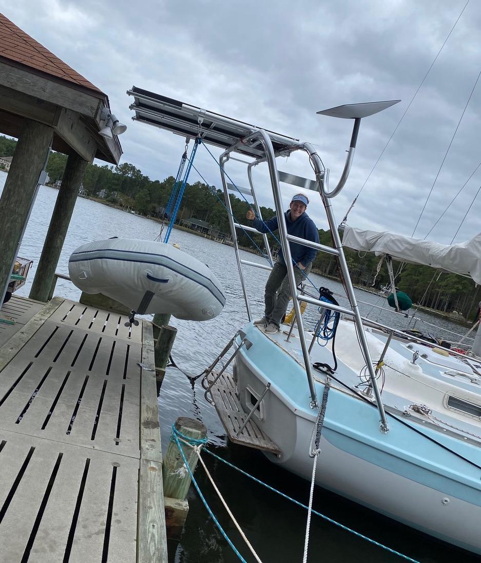 Person standing on a Morgan sailboat adjusting a blue rope attached to an Atlantic Towers Arch lifting a dinghy near a wooden dock.