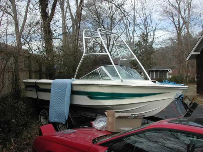 Correct Craft Ski Nautique boat with installed Atlantic Towers Air Tower parked in a wooded outdoor area near a red car.