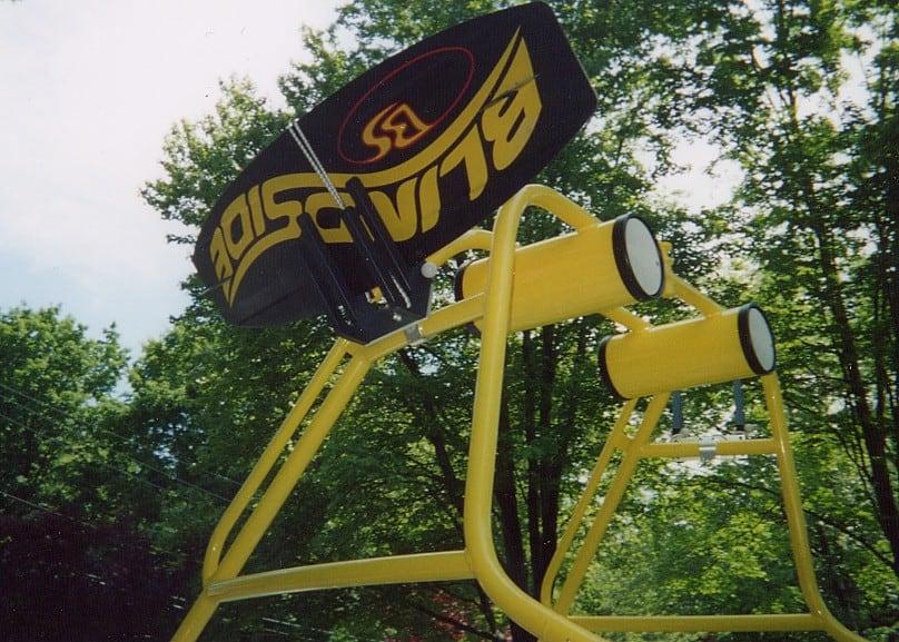Atlantic Towers Accessories mounted on a yellow frame, featuring a large black wakeboard with a logo, amidst greenery.