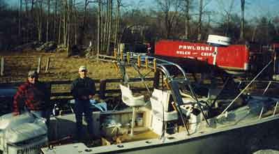 Two men stand on the deck of a Mako 258 Walkaround boat featuring an Atlantic Towers Arch, with trees in the background.