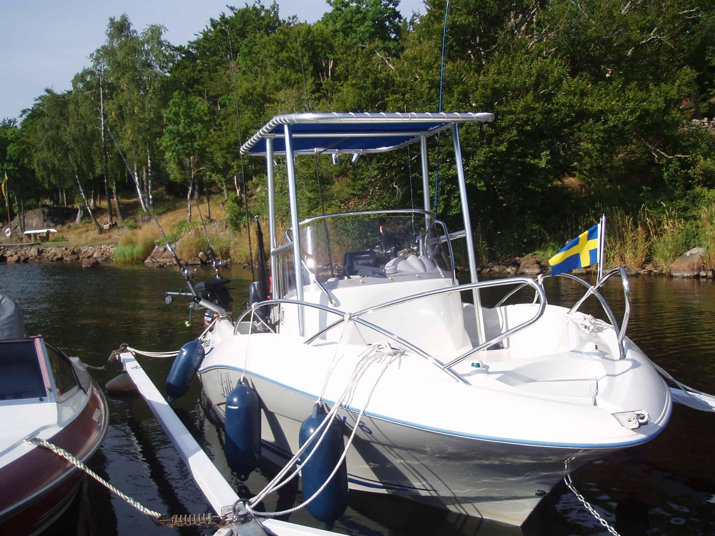 Quicksilver Commander open boat docked with Atlantic Towers T-Top, fishing rods, and Swedish flag on water edge.