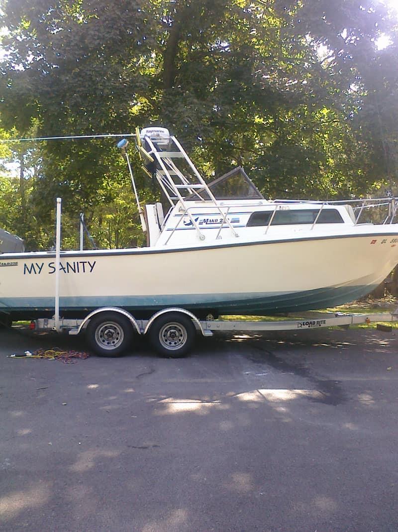 Side view of a Mako 238 W/A boat named My Sanity on a trailer equipped with an Atlantic Towers Arch, parked outdoors near trees.