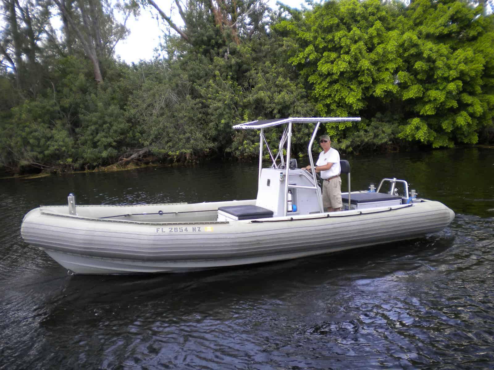 Man standing at the helm of a Willard boat outfitted with an Atlantic Towers T-Top, floating on calm water near dense trees.